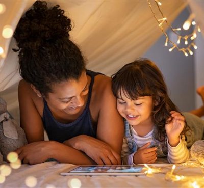 African mother and cute smiling girl using digital tablet while lying in illuminted tent in kid bedroom. Cheerful ethnic woman and lovely daughter on video call under a cozy hut. Lovely little girl with mom watching cartoon on digital tablet in bedroo